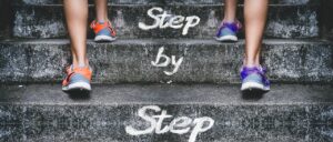 階段を一歩ずつ登る人の足元と “Step by Step” の文字