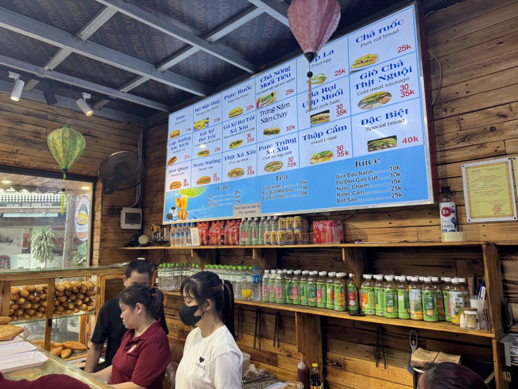 Interior of a Vietnamese banh mi shop showing menu board, drinks, and staff preparing food